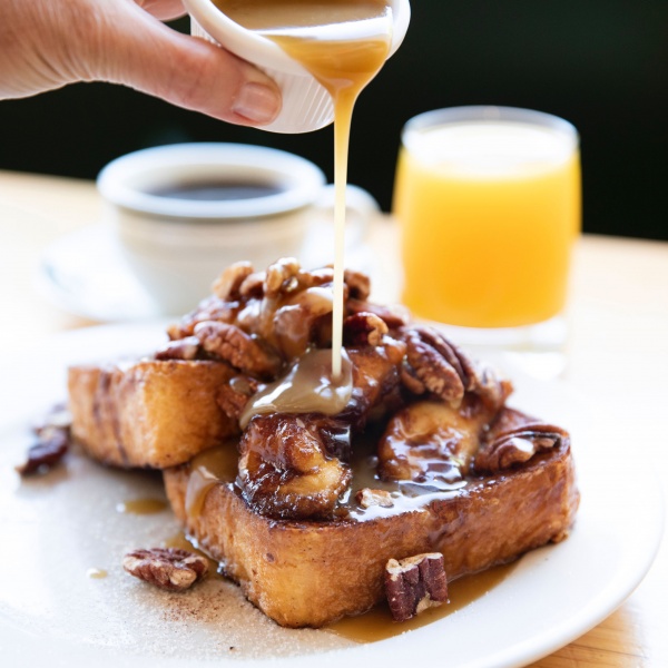 Brioche French Toast with bananas and roasted pecans with maple butter poured on top. Black coffee and OJ in the background.