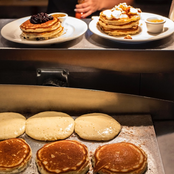 A pancake griddle with cooked pancakes, uncooked pancakes and two stacks of pancakes, blueberry and banana walnut.